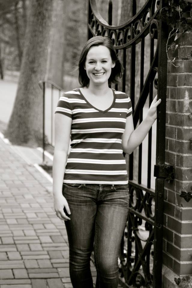 Senior portrait of me (Leah Tams). It is a black and white photo, and I'm standing at an iron gate next to a brick wall on Mary Washington's campus.